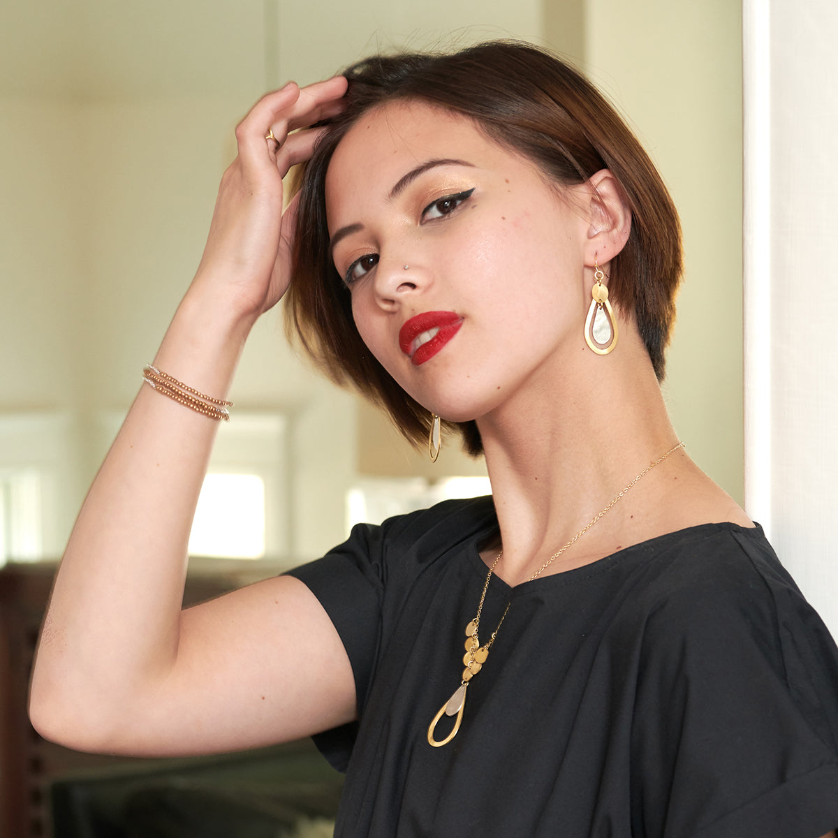 A woman wearing a black shirt and bright red lipstick, looking confidently at the camera, modeling a stack of three bracelets on her arm.