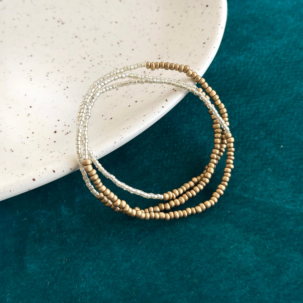 A stack of three gold and silver beaded bracelets rests on a blue cloth against a white plate.