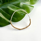 A single silver and gold beaded bracelet rests against a green leaf on a bright white background.