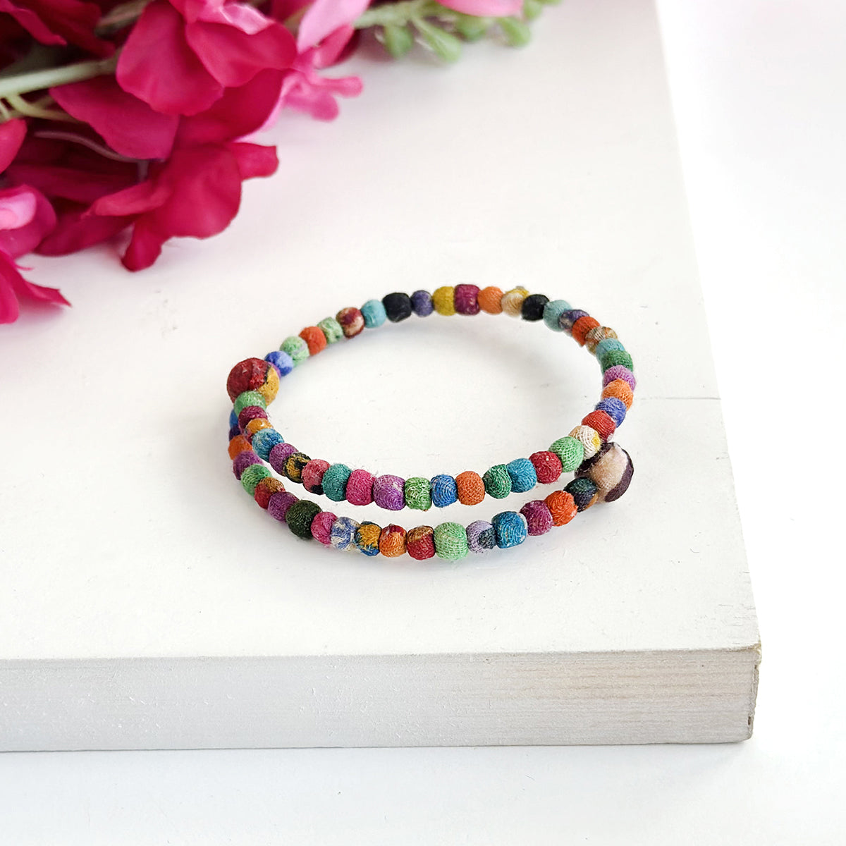 A multicolor beaded bangle bracelet sits on a white wooden board with pink flowers in the background.