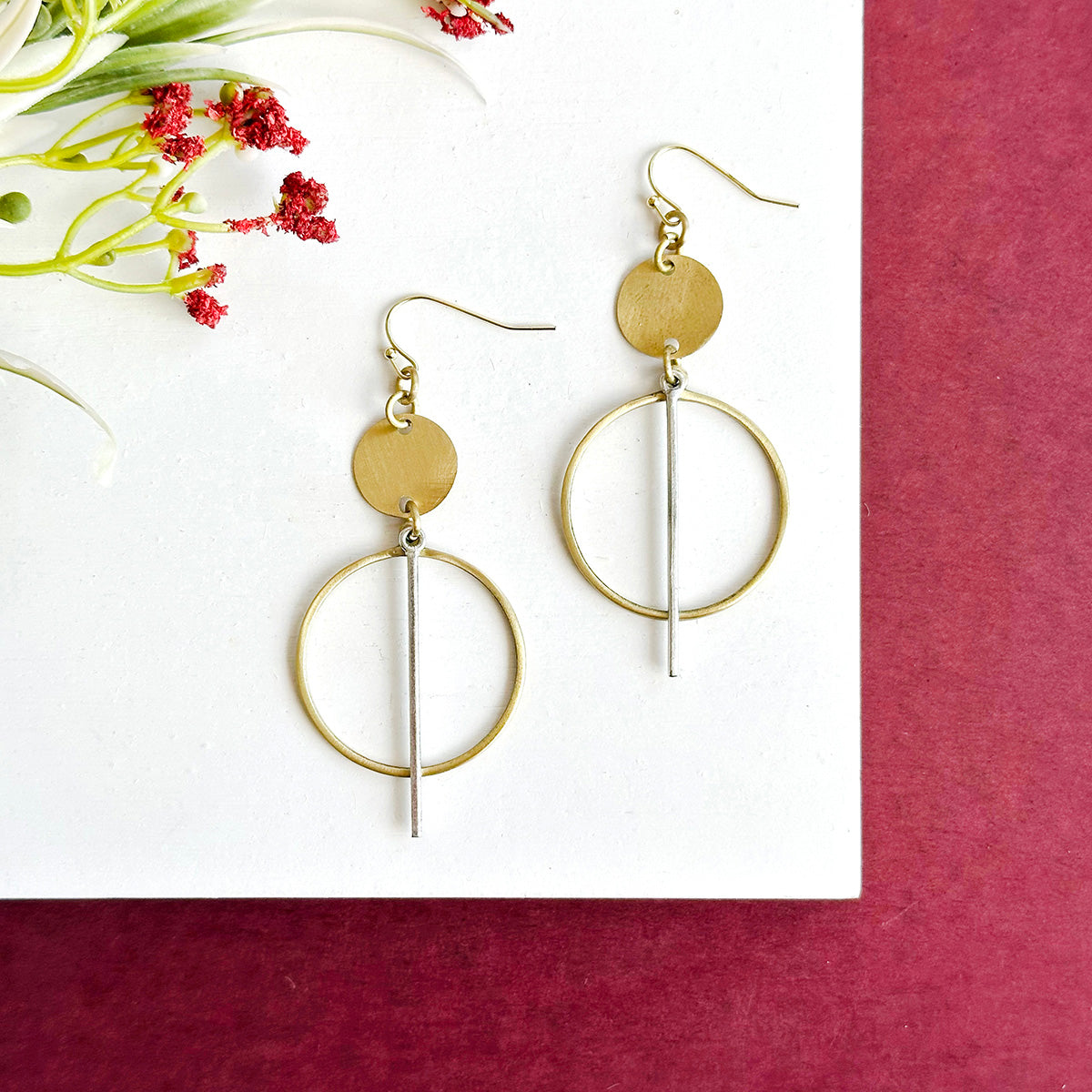 A pair of gold and silver circular earrings rest on a white board with a red background.