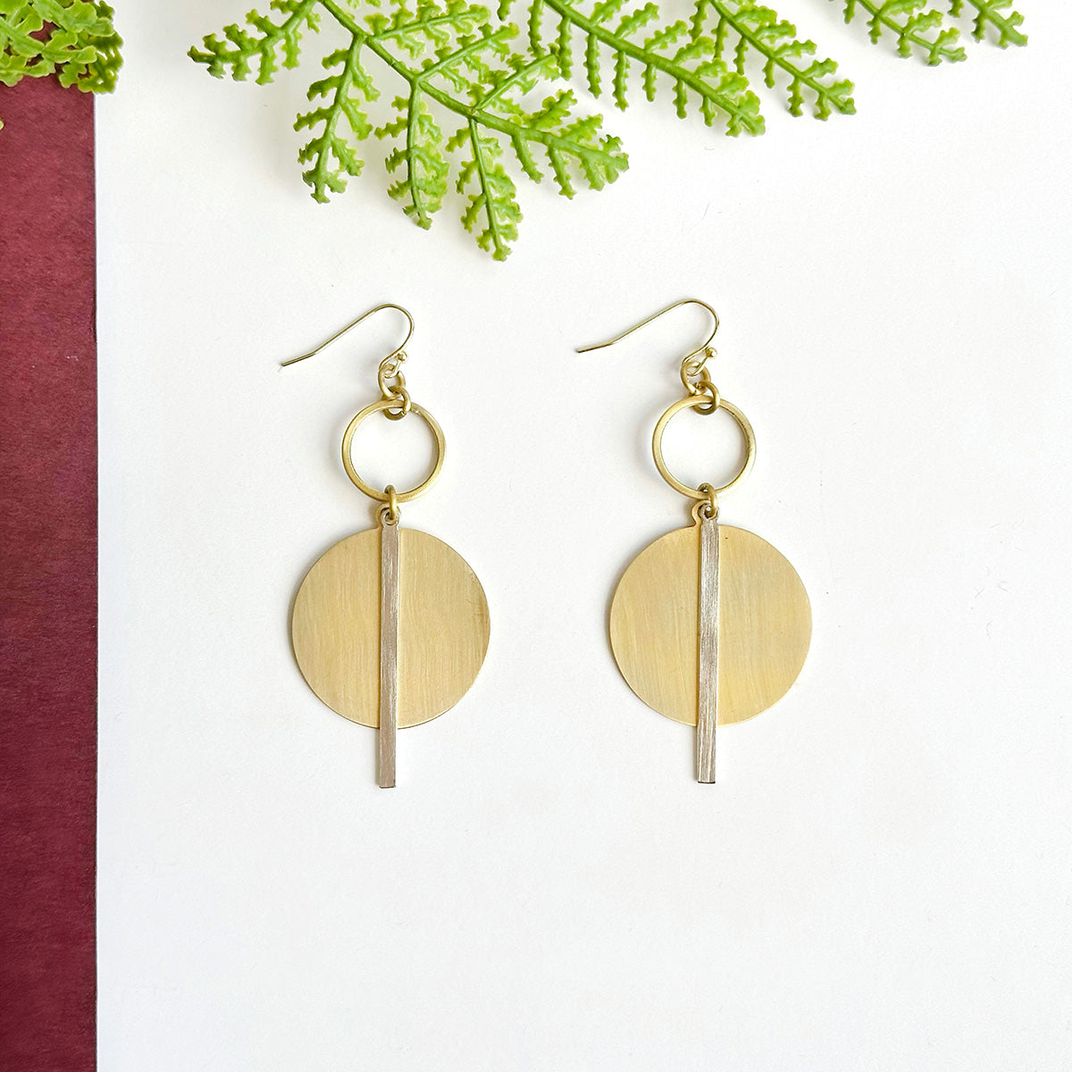 A pair of gold and silver disc earrings rest on a white surface with a green leaf in the background.