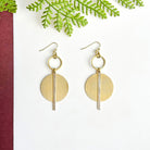 A pair of gold and silver disc earrings rest on a white surface with a green leaf in the background.