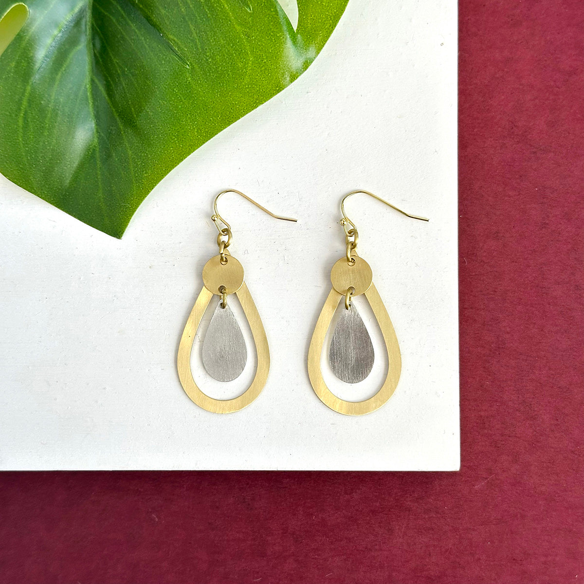 A pair of gold and silver teardrop earrings rests on a white board with a red background and a green leaf.