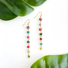 A pair of multicolor, beaded earrings rests against a white surface and is flanked with two green leaves.