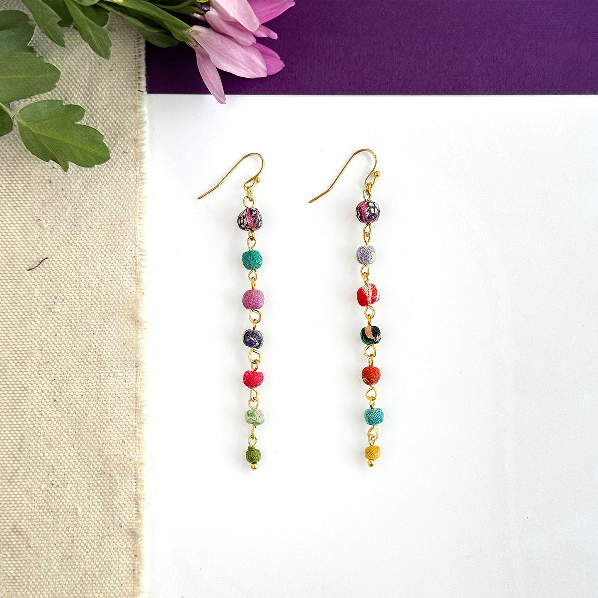 A pair of multicolor beaded earrings rests against a white background with a purple flower in the left.