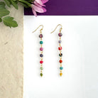 A pair of multicolor beaded earrings rests against a white background with a purple flower in the left.