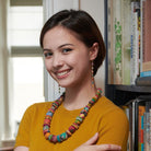 A young woman in a yellow shirt smiles while showing off her multicolor, beaded jewelry.