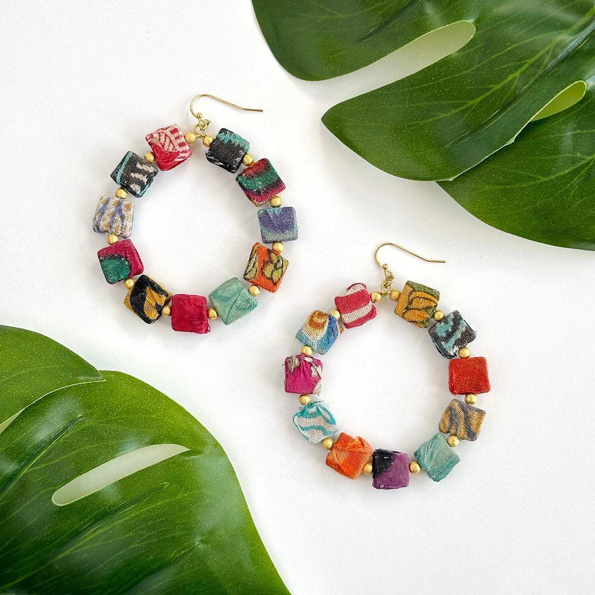 A pair of multicolor, square-beaded hoops is shown against a white surface and flanked by large green leaves.