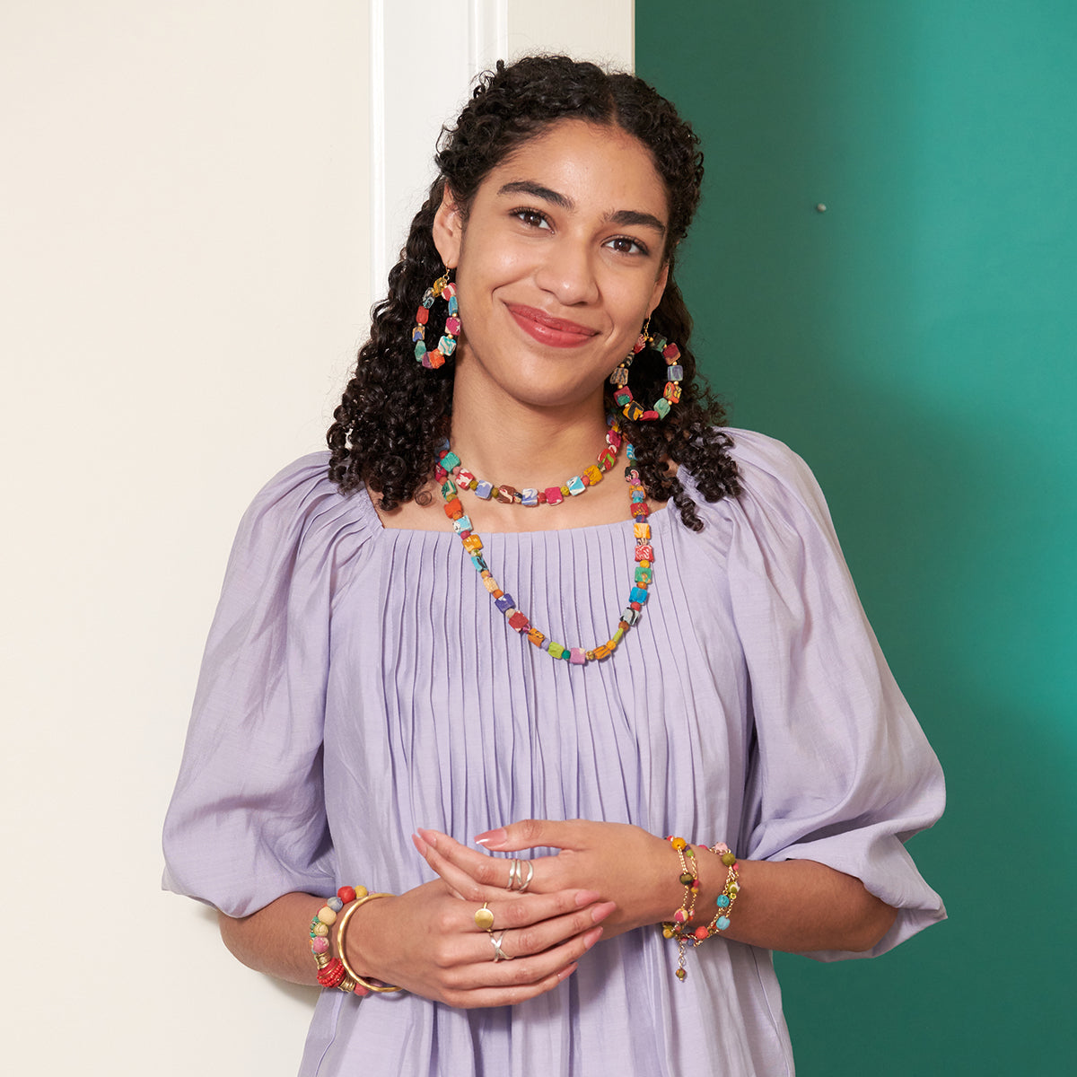 A young woman in a lavender shirt smiles while showing off her multicolor, beaded jewelry.