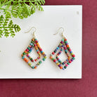 A pair of multicolor beaded earrings rests on a white board against a red background, with a green leaf in the corner.