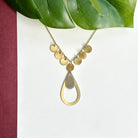 A shiny gold teardrop and charm necklace is shown against a white backdrop with a large green leaf hanging above it.