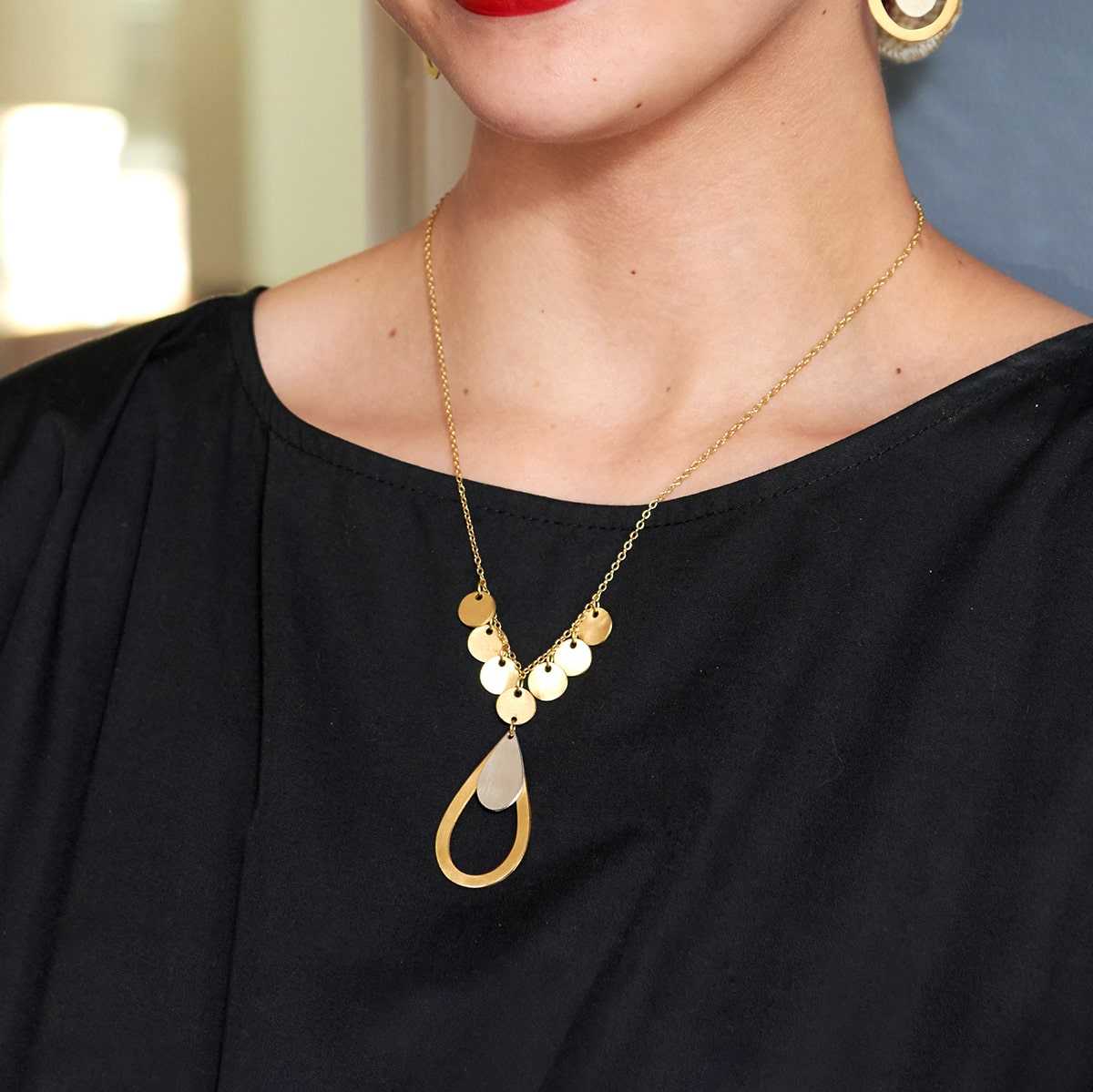 A woman wearing a black dress poses, modeling the gold charm & pendant necklace she is wearing.