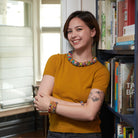 A woman in a yellow top smiles while resting against a bookshelf and modeling multicolored beaded jewelry.