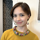 A close up of a woman in a yellow top smiling and modeling her multicolored beaded jewelry.