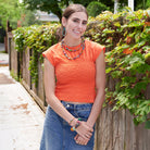 A woman in an orange shirt relaxes outside while crossing her arms. She is adorned with multicolor beaded jewelry.