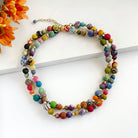 A long multicolor beaded necklace is wrapped twice to show it can be layered. It rests against a white board on a white background with orange flowers in the corner.