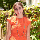 A woman in an orange top stands outside and smiles while modeling multicolor beaded jewelry.