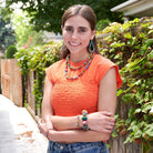 A woman in an orange shirt relaxes outside while crossing her arms. She is adorned with multicolor beaded jewelry.