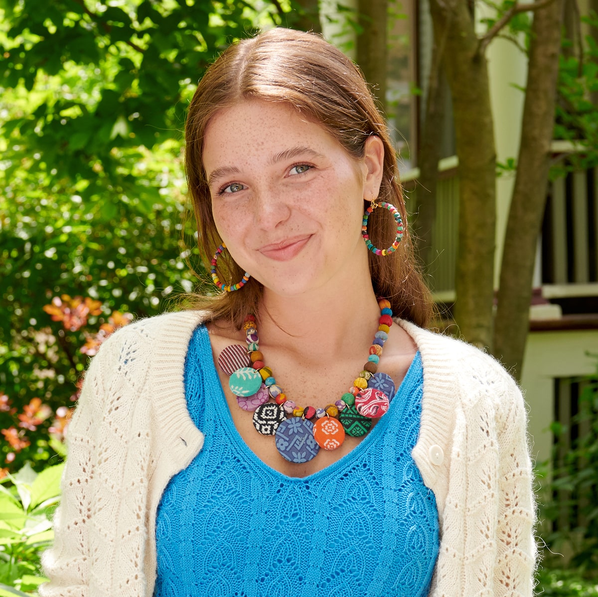 A young woman stands outdoors, wearing a cream cardigan over a blue dress. She is accessorized with colorful beaded jewelry, including a necklace, earrings, and bracelets.