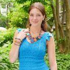 A young woman stands outdoors, wearing a blue dress. She is accessorized with colorful beaded jewelry, including a necklace, earrings, and bracelets.