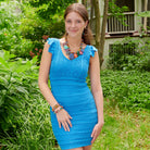 A young woman stands outdoors, wearing a bright blue dress. She is accessorized with colorful beaded jewelry, including a necklace, earrings, and bracelets.