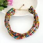 A multicolored and multistrand necklace rests against a white plate, accented by a pink flower in the corner.