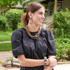 A young woman stands outdoors and smiles, wearing a black dress. She is accessorized with colorful beaded jewelry, including a necklace, earrings, and bracelets.