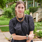 A young woman stands outdoors and smiles, wearing a black dress. She is accessorized with colorful beaded jewelry, including a necklace, earrings, and bracelets.