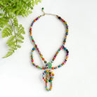 A multicolored beaded necklace rests against a white background, accented by green leaves in the corner.