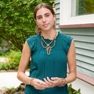 A young woman stands outdoors, wearing a teal top. She is accessorized with colorful beaded jewelry, including a necklace, earrings, and bracelets.