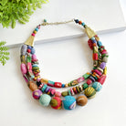 A multicolored beaded necklace rests against a white background, accented by green leaves in the corner.