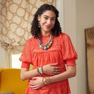 A young woman stands indoors, wearing a bright red top. She is accessorized with colorful beaded jewelry, including a necklace, earrings, and bracelets.