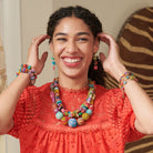 A young woman wearing a bright red top laughs while touching her ears and modeling multicolor beaded jewelry.