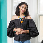 A young woman stands indoors, wearing a black top. She is accessorized with colorful beaded jewelry, including a necklace, earrings, and bracelets.