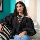 A young woman wearing a black top and jeans sits on a bench by a window. She is accessorized with colorful beaded jewelry, including a necklace, earrings, and bracelets.