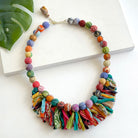 A multicolored beaded & fringe necklace rests against a white board, accented by a green leaf in the corner.