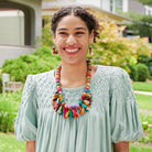 A young woman stands outdoors and laughs, wearing a light blue top. She is accessorized with colorful beaded jewelry, including a necklace, earrings, and bracelets.