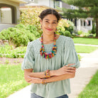 A young woman stands outdoors and smiles, wearing a light blue top. She is accessorized with colorful beaded jewelry, including a necklace, earrings, and bracelets.