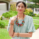 A young woman stands outdoors and smiles, wearing a light blue top. She is accessorized with colorful beaded jewelry, including a necklace, earrings, and bracelets.