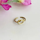 A gold ring, with a center pretzel design, rest against a white surface with pink flowers in the background.