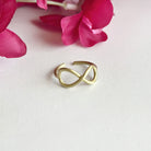 A gold ring with center infinity sign motifs sits against a white surface with a red flower in the background.