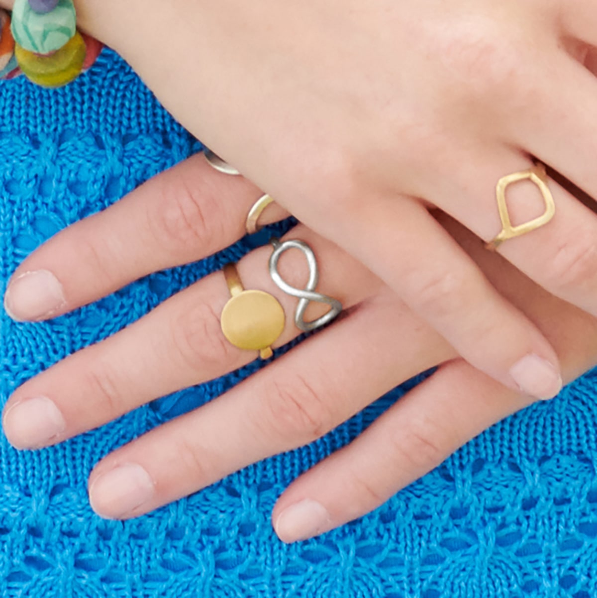 A woman wearing a blue dress holds her hands to her side, showing off the many gold rings on her fingers.