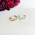 Gold and silver rings with center infinity sign motifs sit against a white surface with a red flower in the background.