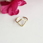 A gold ring with a cutout rectangular center motif rests on a white background with a pink flower in the background.