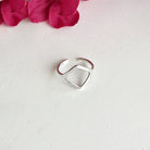 A silver ring with a cutout rectangular center motif rests on a white background with a pink flower in the background.