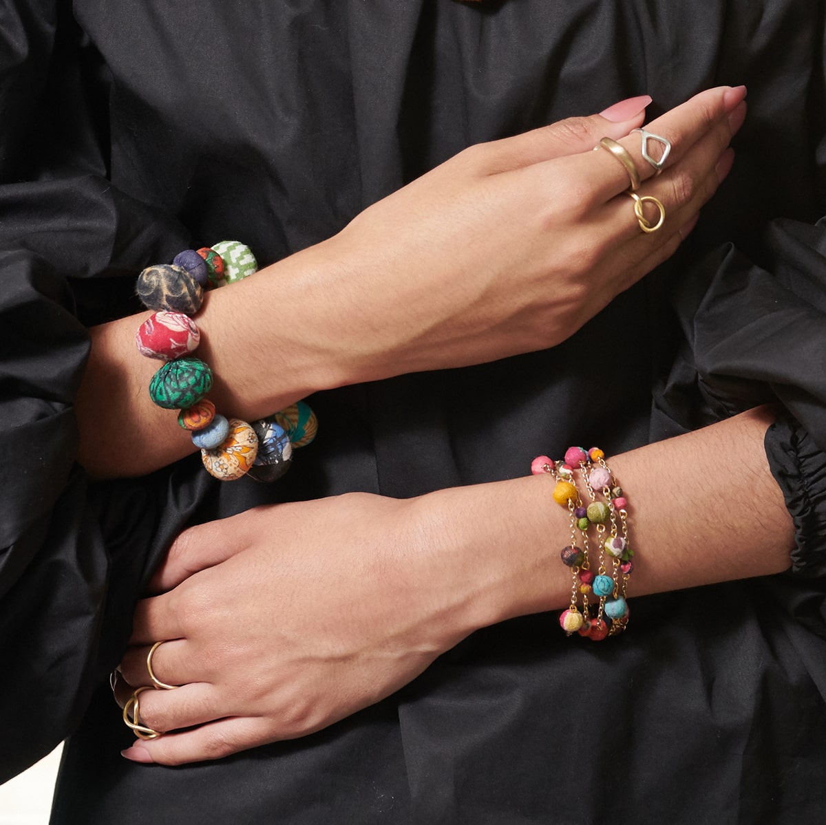 A woman in a black dress crosses her hands across her waist. She is accessorized by many gold and silver rings, and a stack of multicolor beaded bracelets.