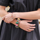 A woman in a black dress crosses her hands across her waist. She is accessorized by many gold and silver rings, and a stack of multicolor beaded bracelets.