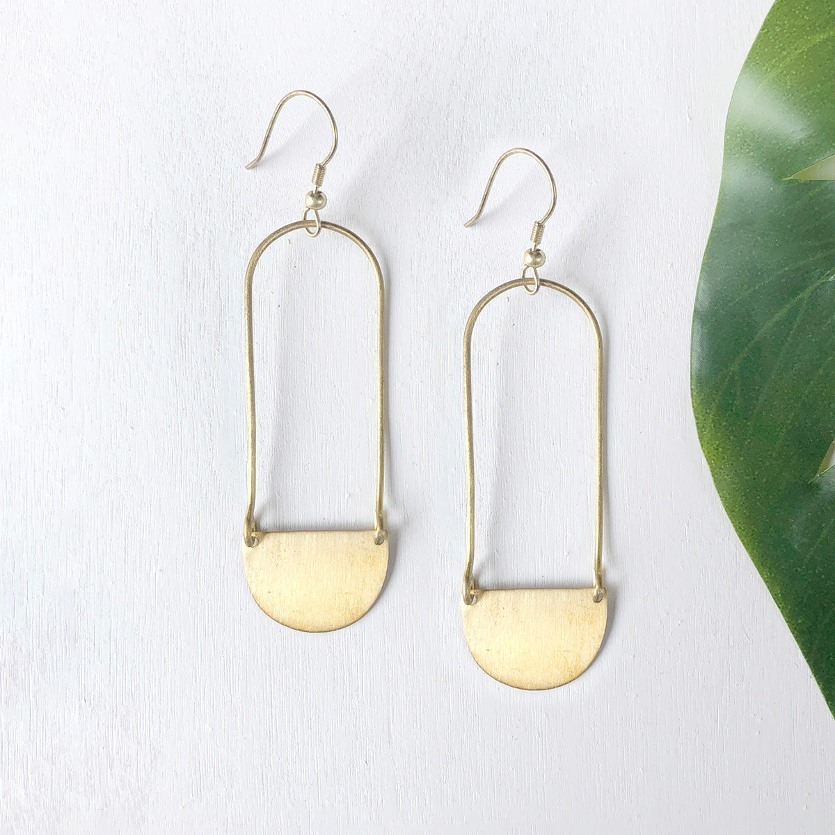 A pair of gold dangling earrings against a white background with a green leaf on the right.