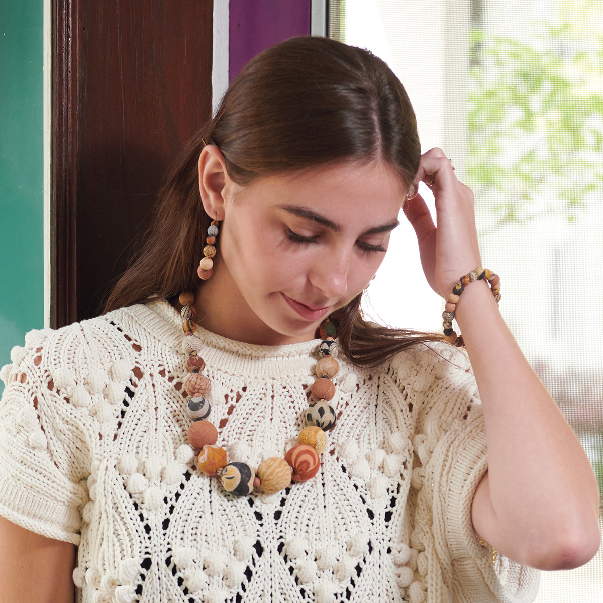 A woman wearing a tan shirt and beaded jewelry scratches her head and looks down.